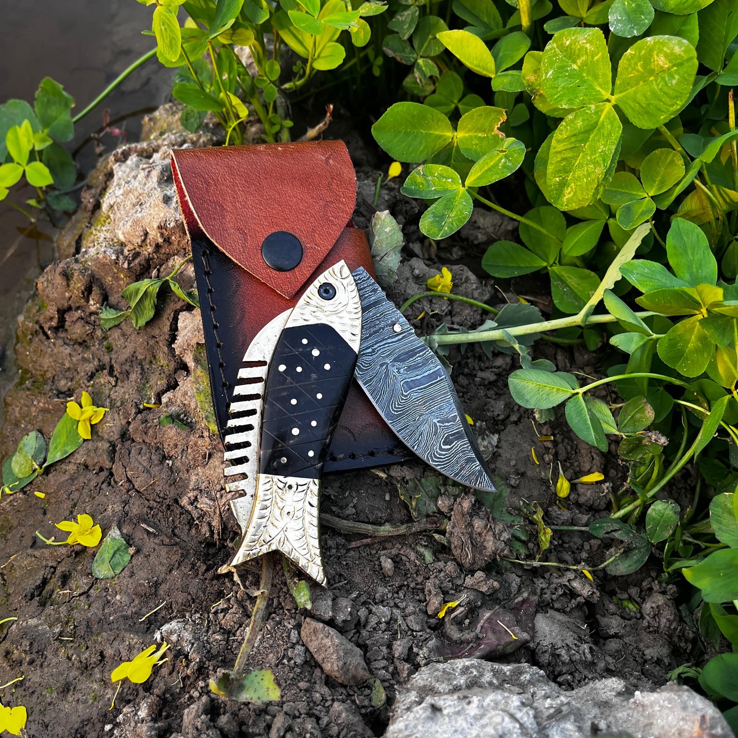 Handmade Damascus folding knife fish design displayed on natural soil background emphasizing outdoor ready everyday carry knife.
