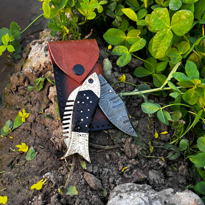 Handmade Damascus folding knife fish design displayed on natural soil background emphasizing outdoor ready everyday carry knife.