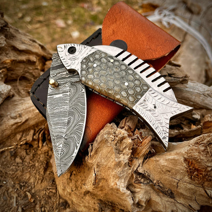 Damascus fish pocket knife partially open showing patterned steel blade and decorative fish handle design in outdoor setting.