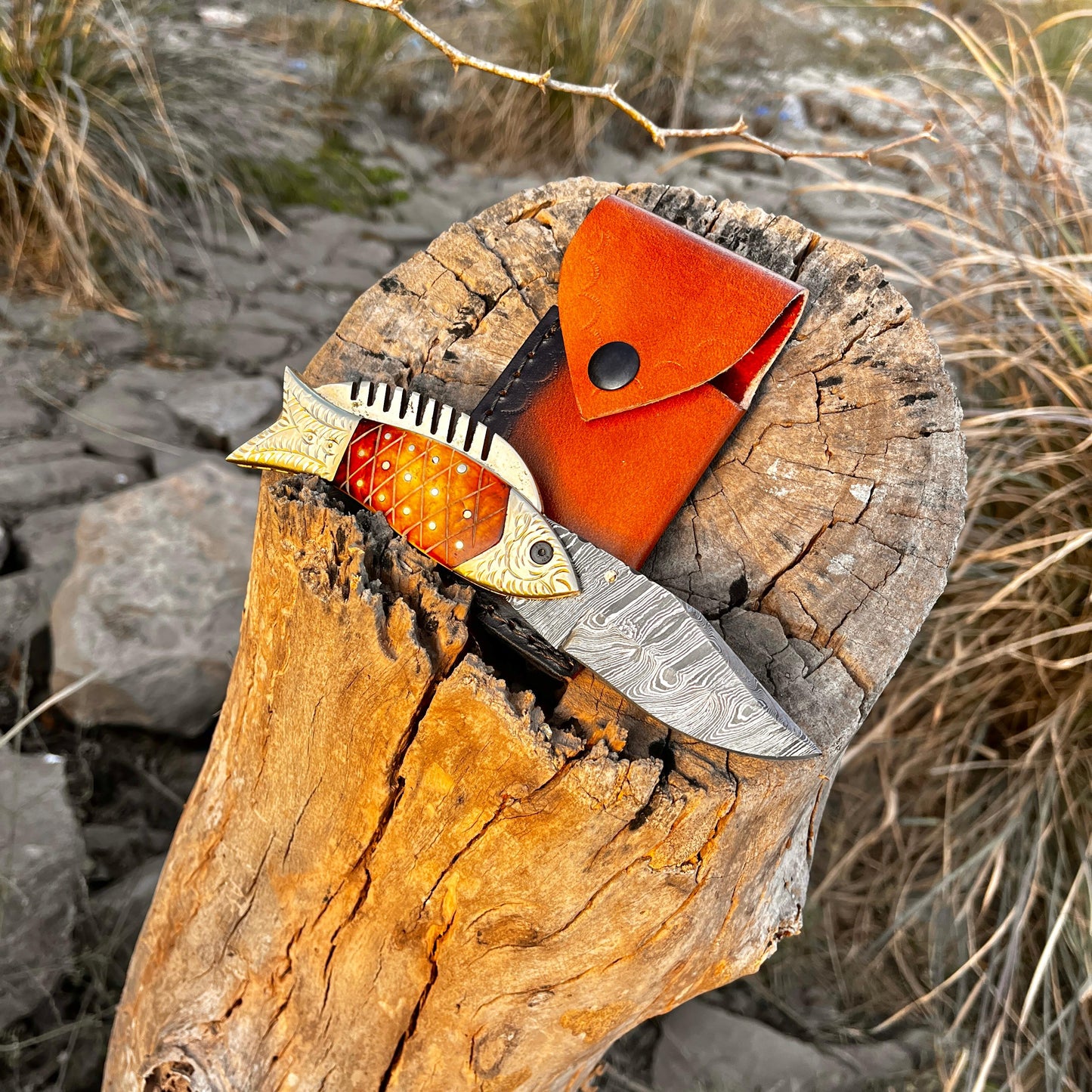 Handmade Damascus fish pocket knife displayed on tree stump with leather sheath showing outdoor ready design and durable folding mechanism.