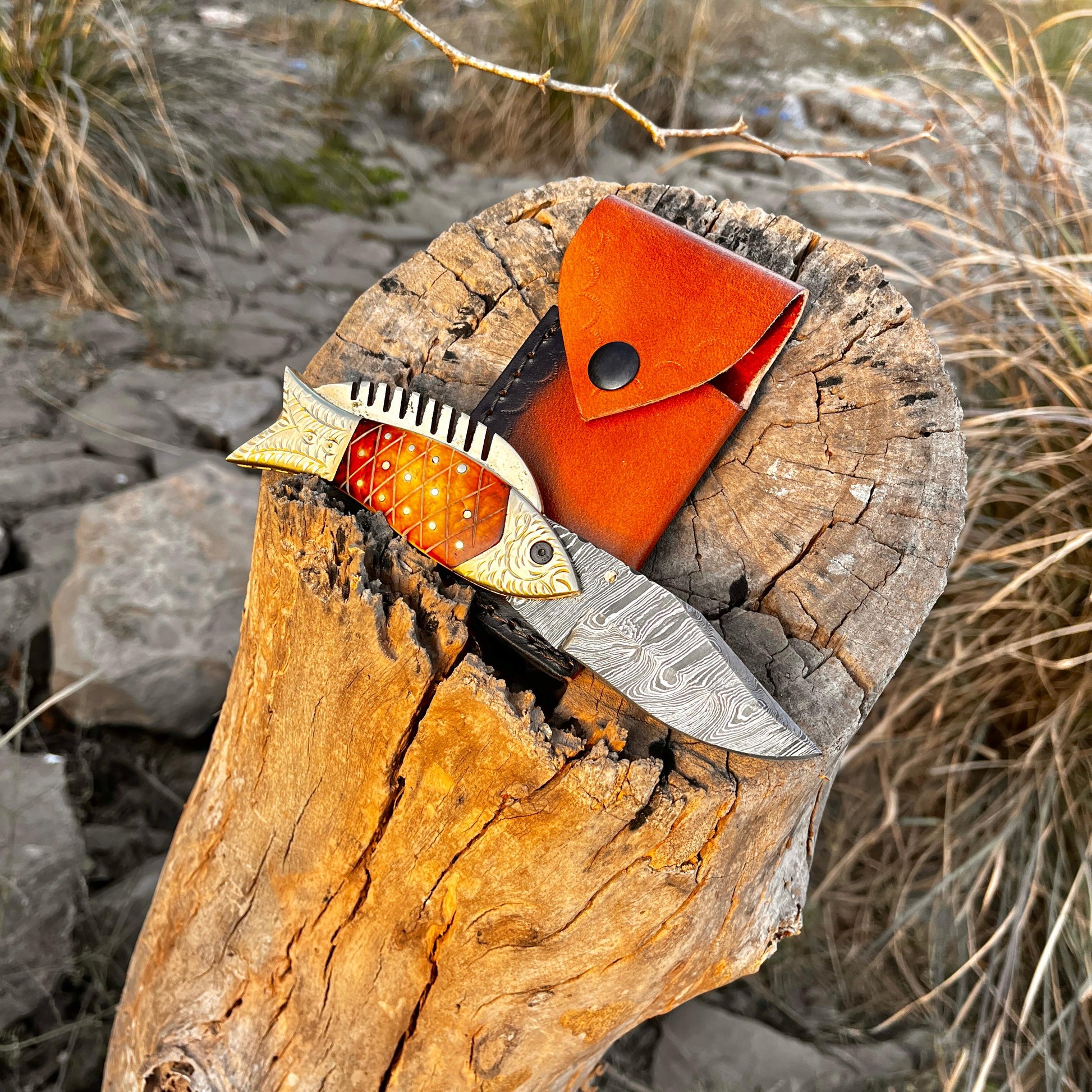 Handmade Damascus fish pocket knife displayed on tree stump with leather sheath showing outdoor ready design and durable folding mechanism.