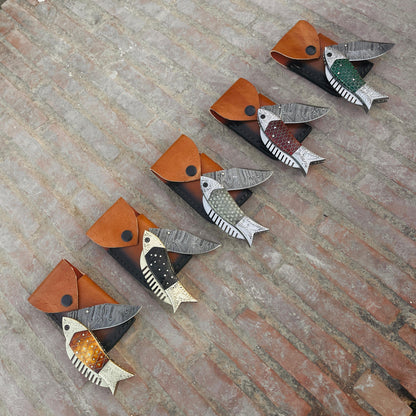 Handmade fish Damascus folding pocket knife displayed with leather pouch on outdoor brick background showing detailed Damascus blade pattern.