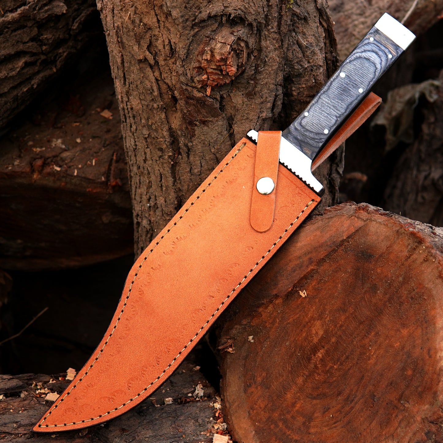 Bowie knife displayed with leather sheath on wooden logs in outdoor environment showing protective carry option and rugged field style presentation. The knife is positioned to demonstrate blade length, sheath quality, and outdoor readiness, making it appealing for camping gear collections and gift buyers searching for a strong and stylish fixed blade knife.