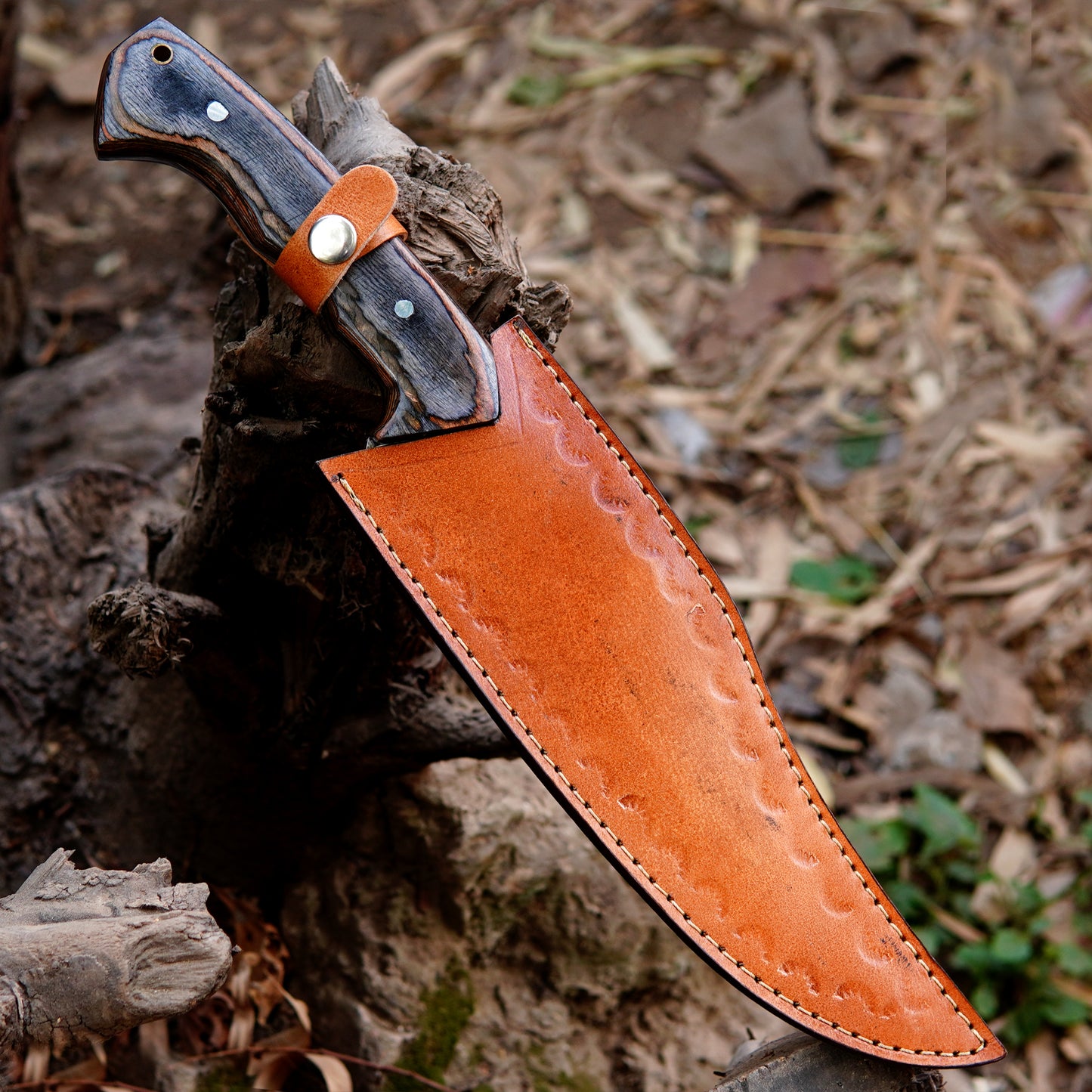 Outdoor display image of a full tang bowie knife resting on wood logs with leather sheath beside it. The polished blade reflects natural light, showing clean grind lines and sharp edge. This image represents the knife as a reliable outdoor companion suitable for camping, survival activities, and collectors who value handcrafted tools.