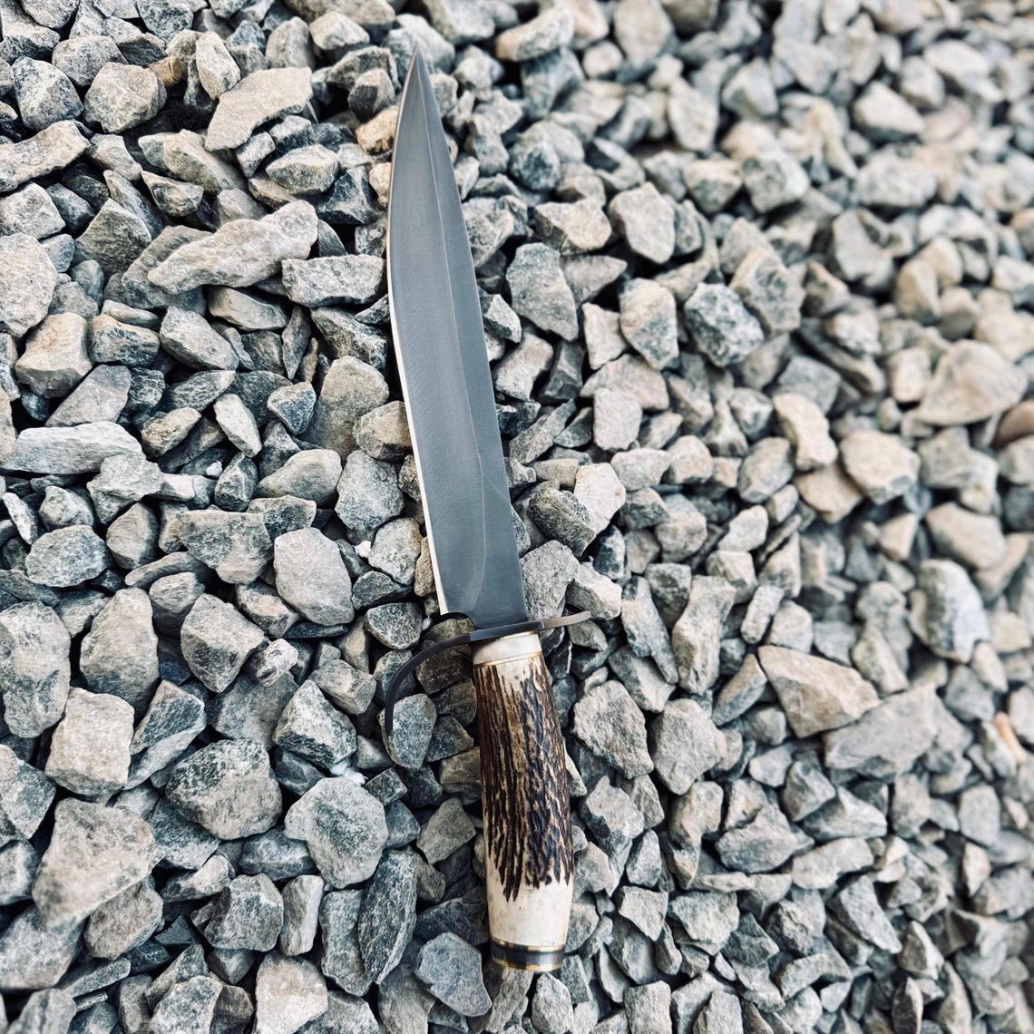 Handmade Bowie knife resting in a black leather sheath with contrast stitching, featuring a strong 5160 spring steel blade and antler-style handle. Built for lasting durability.