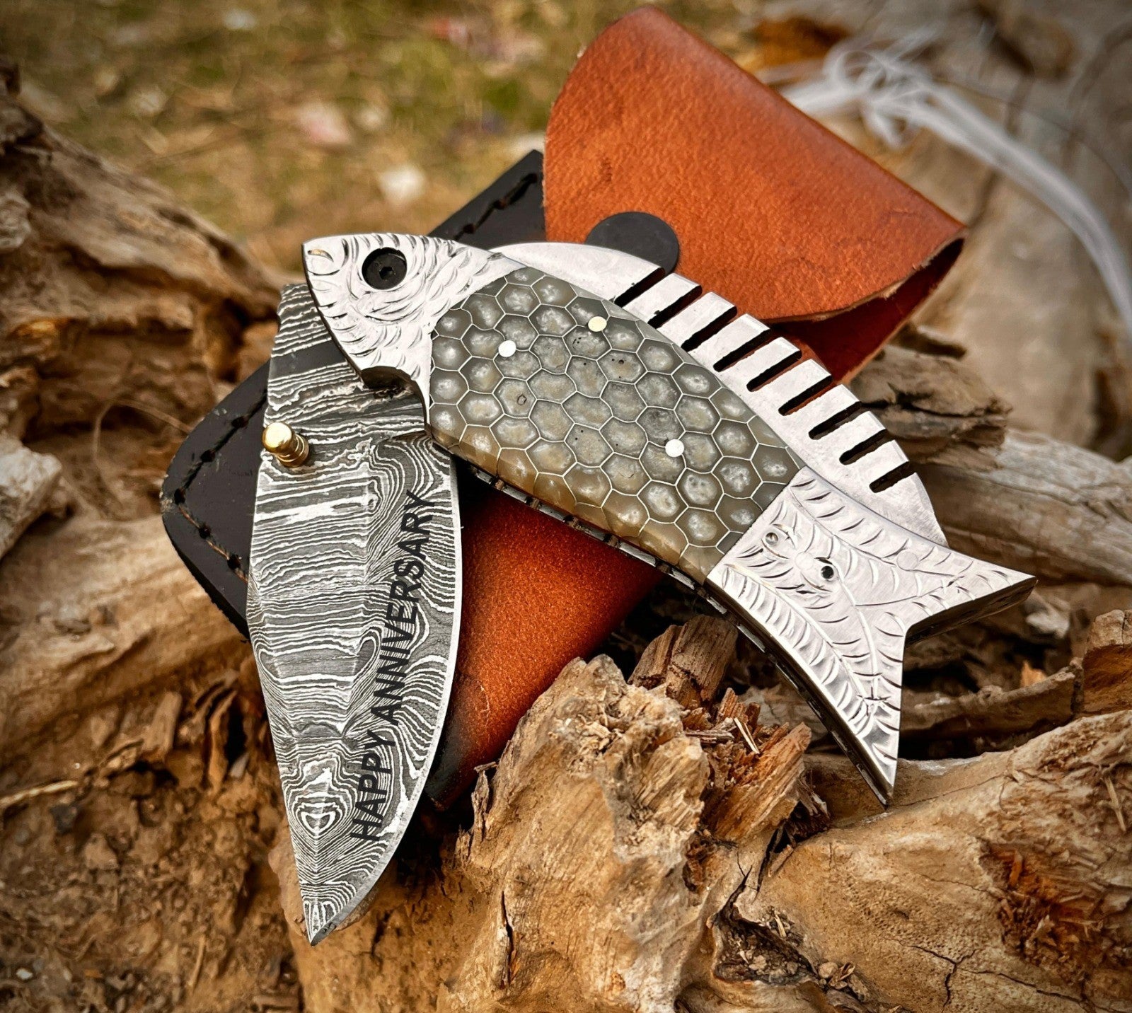 Fish shaped folding knife with personalization and monogram options displayed with leather sheath on wooden background.