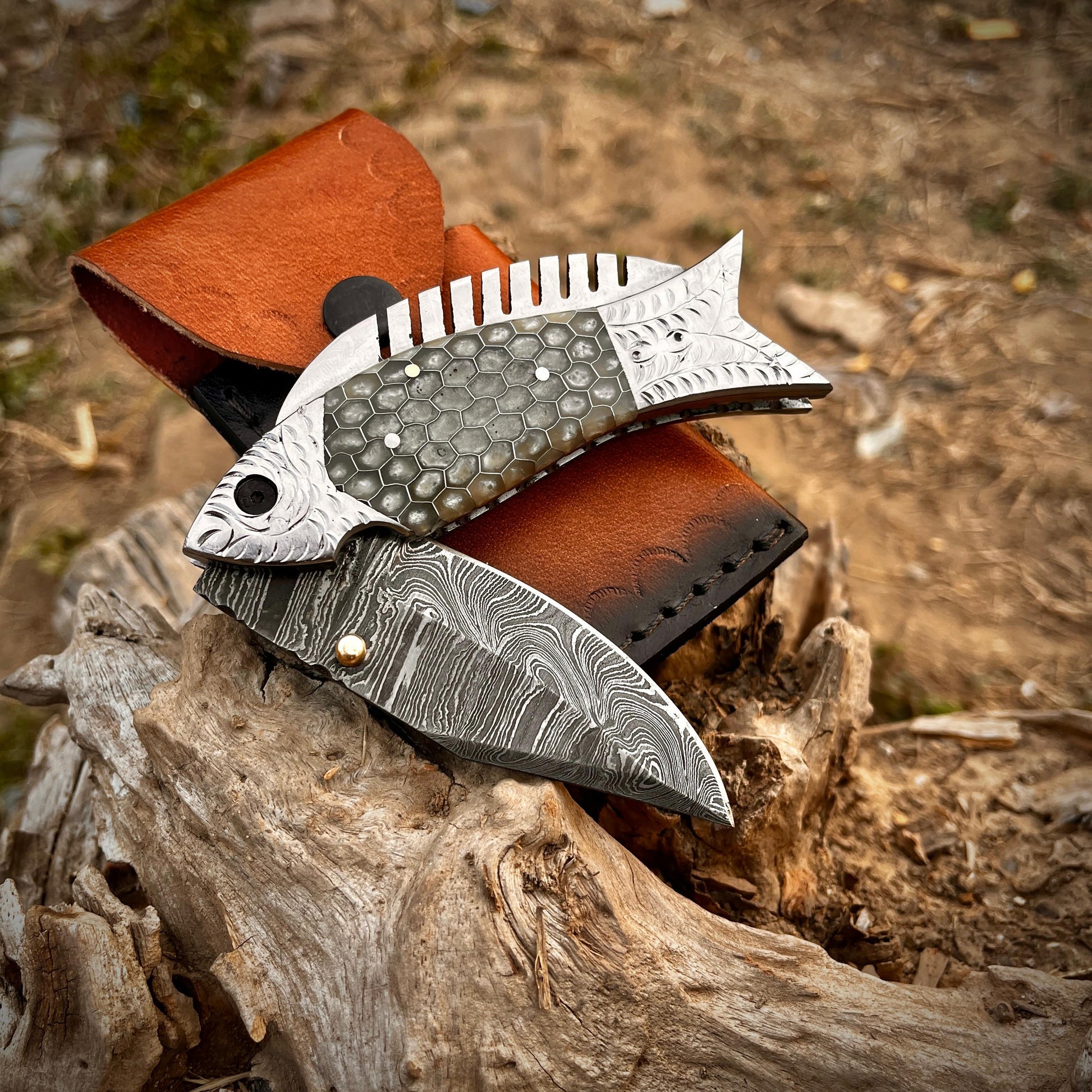 Grey scale fish Damascus folding knife displayed on wooden surface with leather sheath emphasizing craftsmanship and outdoor style.