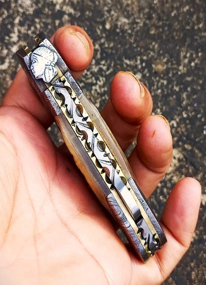 Damascus steel EDC knife with ram horn handle displayed on textured surface.