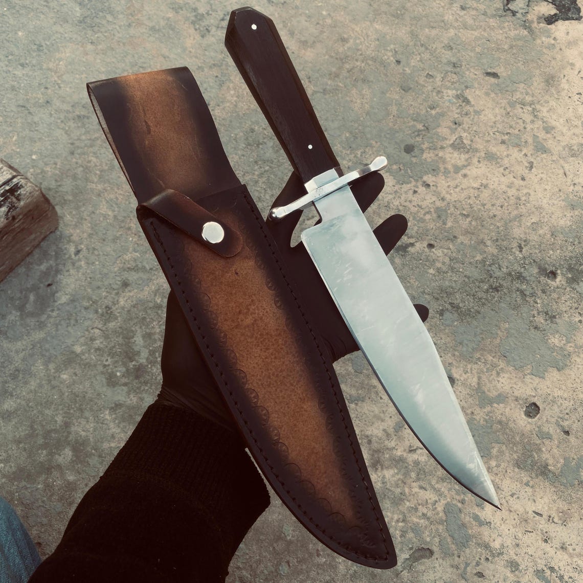 Handmade carbon steel Bowie knife displayed with matching stitched leather sheath.