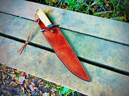 Full-length handmade sandbar bowie knife inside its leather sheath placed outdoors on stone bench.