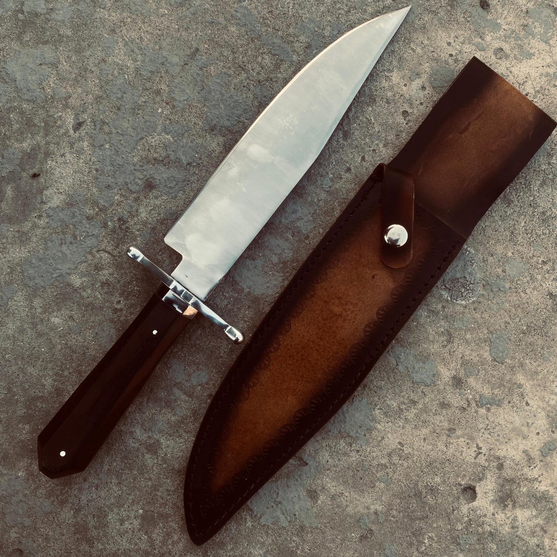 Handmade Bowie knife with long carbon steel blade and stitched leather sheath on stone background.