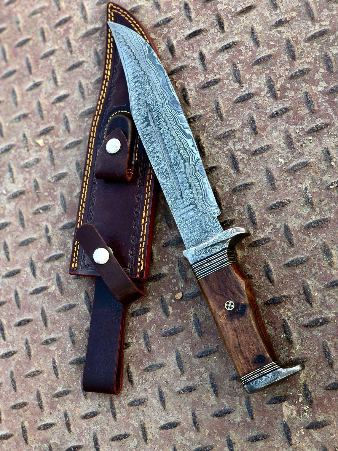Handmade Damascus bowie knife with 9-inch patterned blade and blue pakka wood handle shown on textured background with leather sheath.