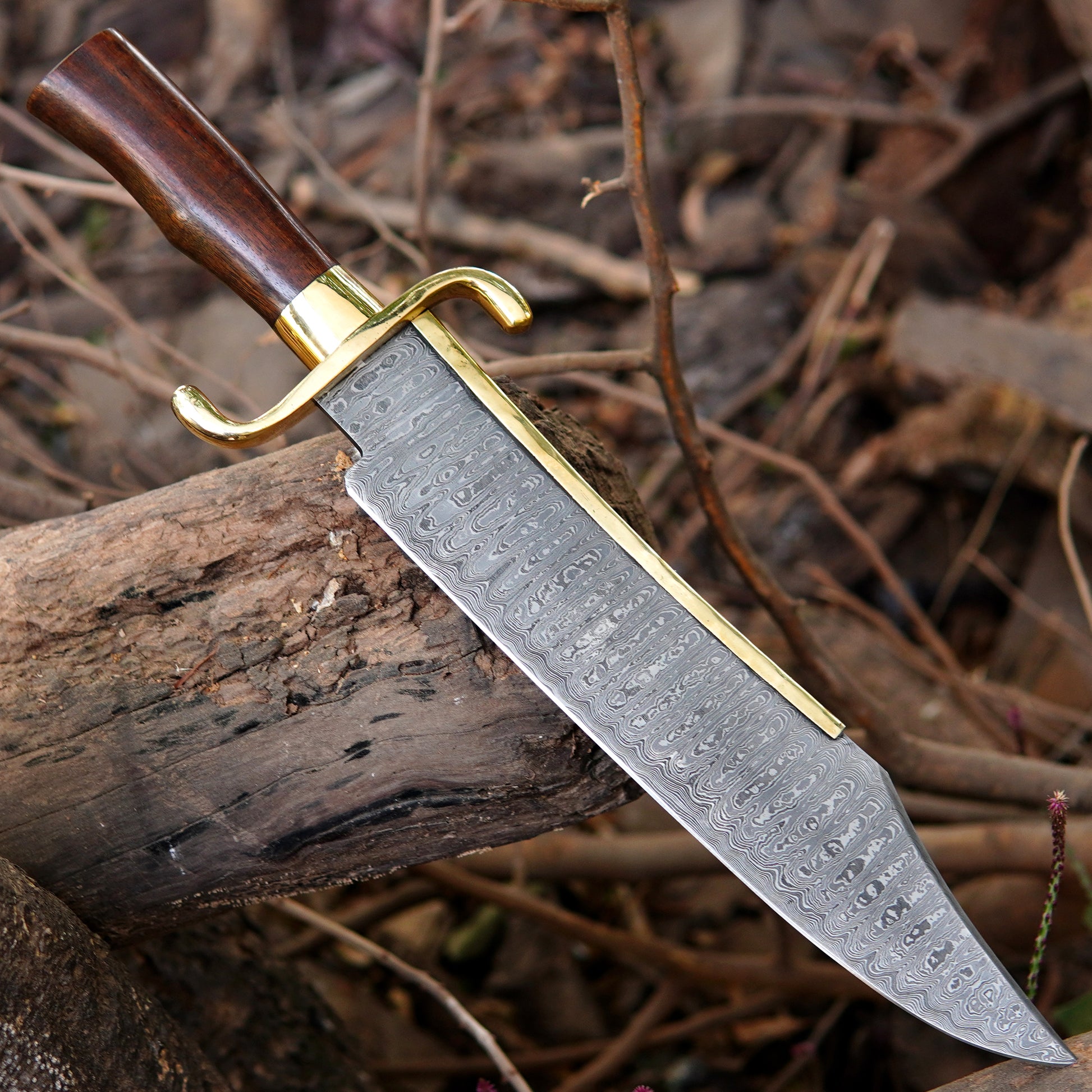 Close view of Damascus steel bowie knife displayed outdoors with focus on blade pattern and overall knife balance. The natural background enhances the handmade appearance while showing the sharp edge and classic shape, making it ideal for product presentation in outdoor gear, handmade tools, and collector knife listings.