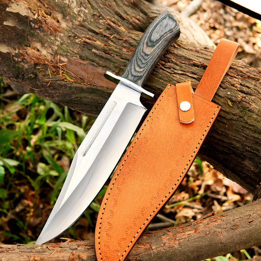 Handmade carbon steel bowie knife placed outdoors on wood with sharp polished blade and grey patterned handle showing strong full tang construction and durable leather sheath designed for camping hunting and survival use