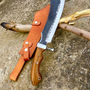 Carbon steel machete placed on natural surface beside handmade leather sheath for outdoor display.