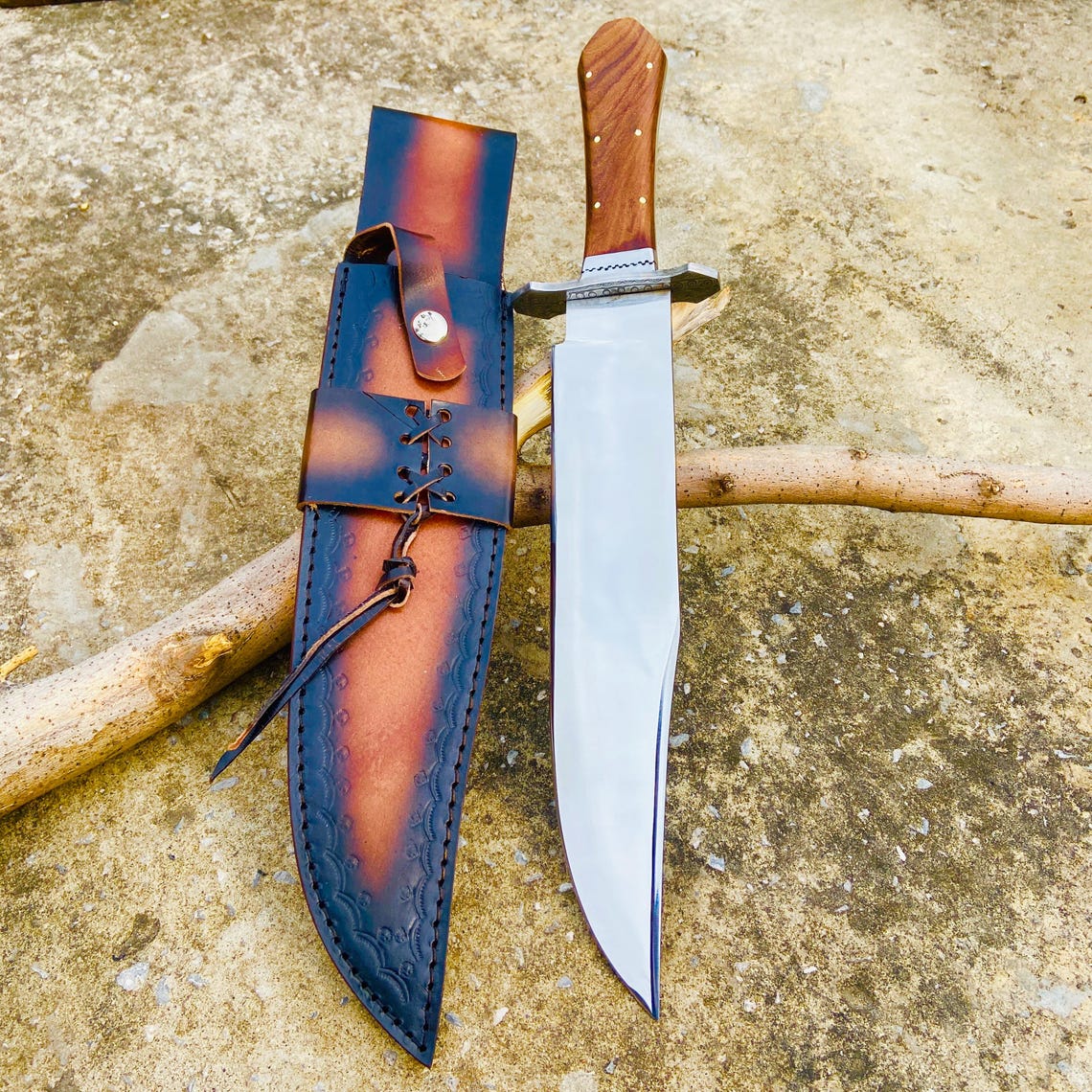 Hand-forged carbon steel Bowie knife displayed beside its premium two-tone leather sheath. The mirror-like blade surface and sharp edge highlight its high craftsmanship. Perfect for hunting, bushcraft, and outdoor activities.