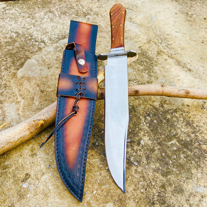 Hand-forged carbon steel Bowie knife displayed beside its premium two-tone leather sheath. The mirror-like blade surface and sharp edge highlight its high craftsmanship. Perfect for hunting, bushcraft, and outdoor activities.