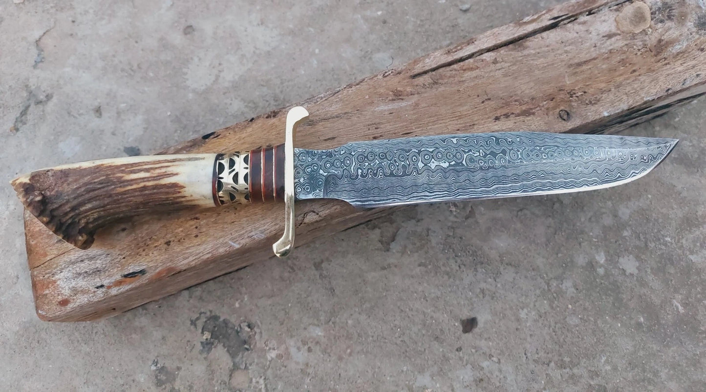 Bowie-style hunting knife crafted in Damascus steel with crown stag handle and brass spacers. Built with a hand-stitched leather sheath for safe carry. A handmade custom knife ideal for hunters, outdoor enthusiasts, and collectors.