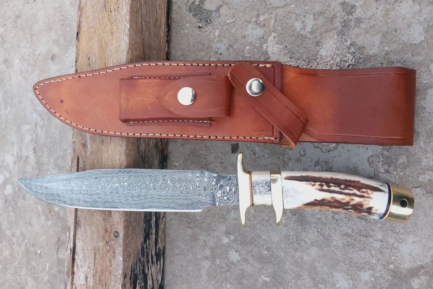 Custom Damascus steel hunting knife with brass guard, stag handle, and sharp patterned blade. Includes a durable hand-stitched sheath. Designed for outdoor use, hunting, and display in collections, balancing artistry with practical use.
