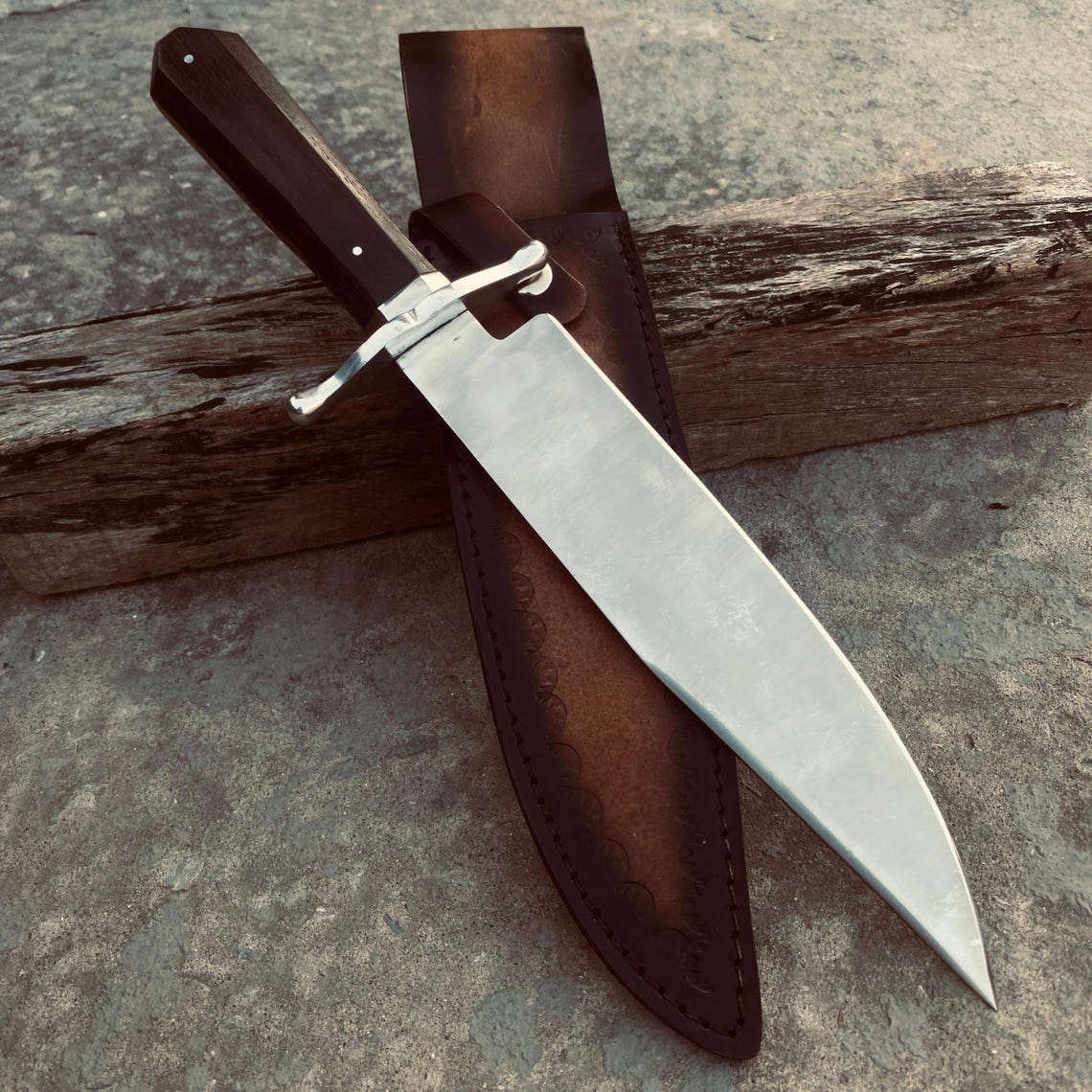 Bowie knife fully inserted in its leather sheath resting on rustic wooden surface.