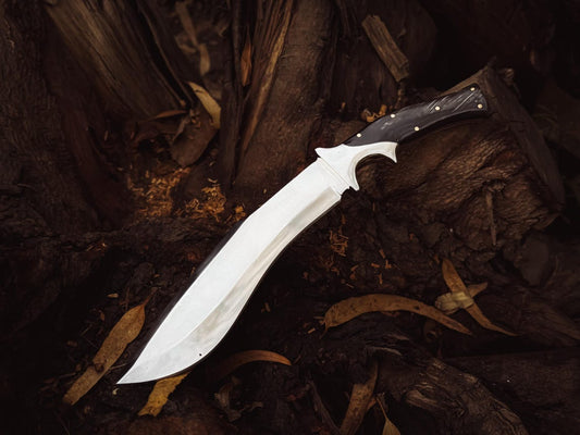 Handmade kukri-style bowie knife standing beside a leather sheath on stacked wood.