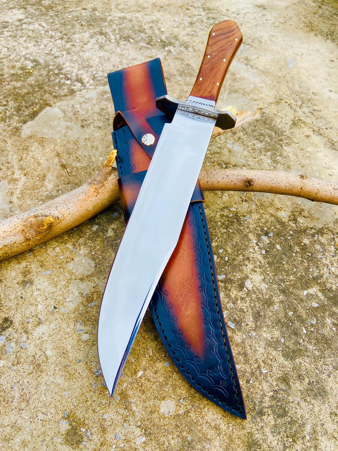 Top view of full-tang handmade carbon steel Bowie knife with rosewood handle and mirror finish blade. The leather sheath beside it shows detailed craftsmanship and secure fit. A collectible handmade knife ideal for outdoor enthusiasts.