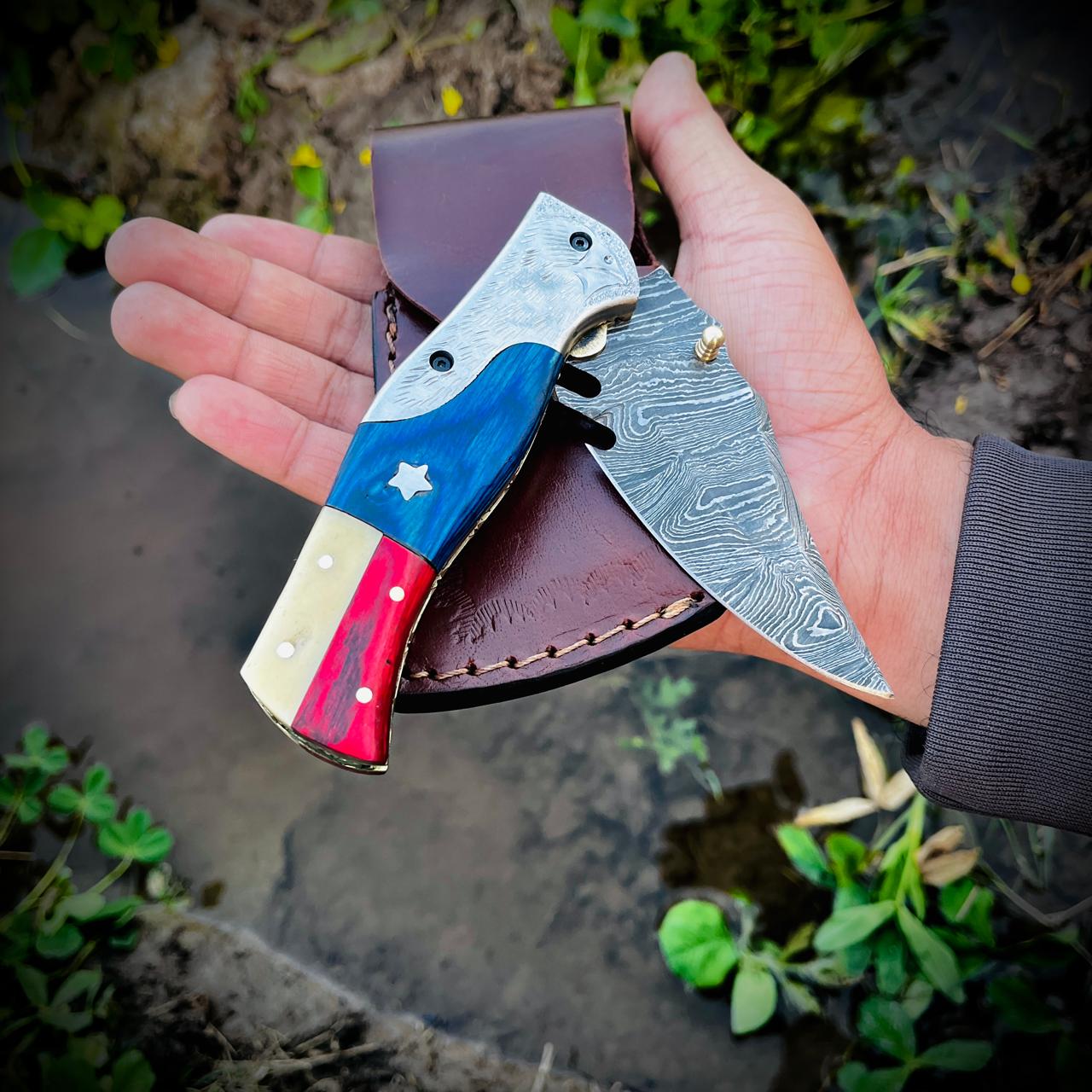 Handmade Damascus steel folding pocket knife with patriotic red white blue handle displayed with leather pouch on natural outdoor background.