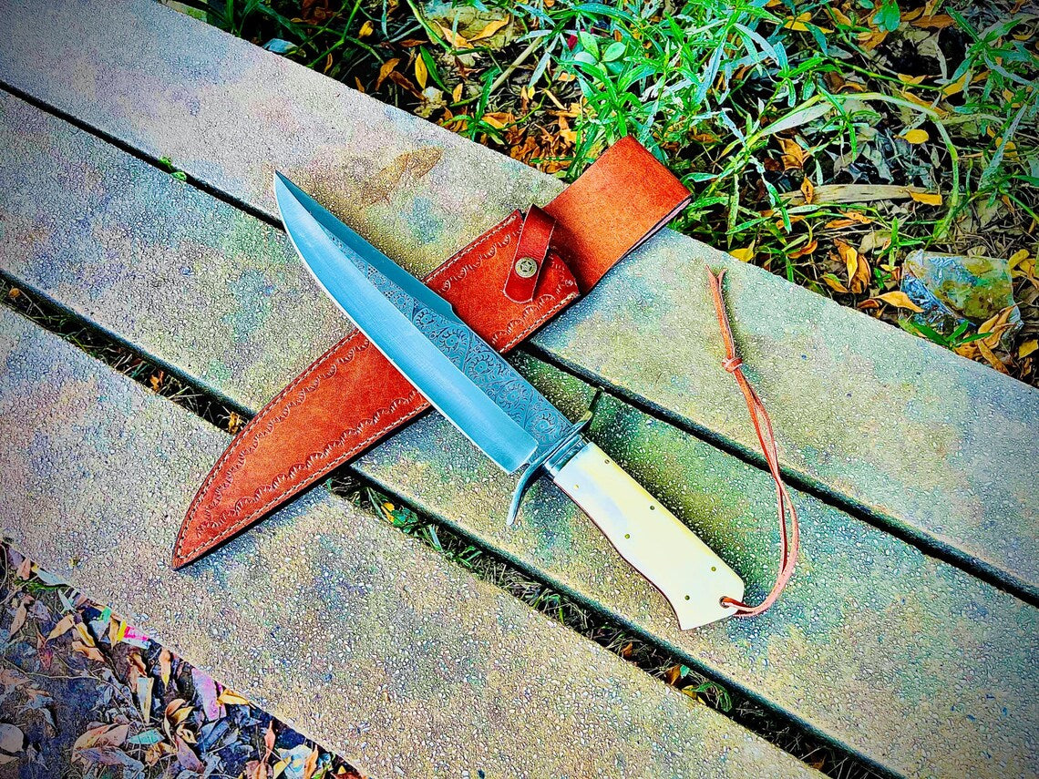 Handmade sandbar bowie knife with engraved carbon steel blade and natural material handle resting beside leather sheath on stone surface.