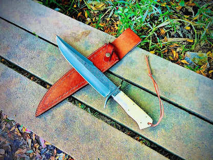 Handmade sandbar bowie knife with engraved carbon steel blade and natural material handle resting beside leather sheath on stone surface.