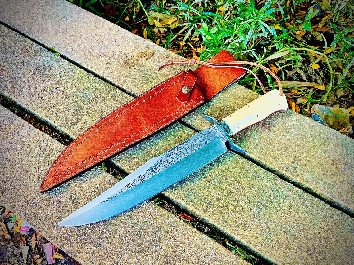 Outdoor photo of engraved carbon steel bowie knife with full tang construction and leather sheath.