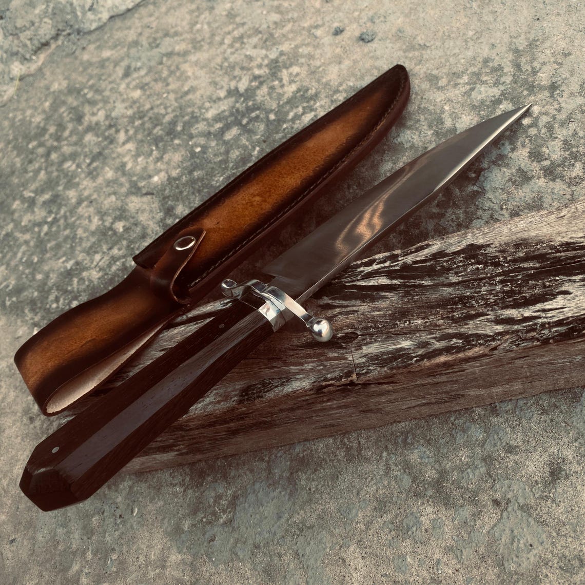 Display shot of carbon steel Bowie knife with leather sheath on textured ground surface.
