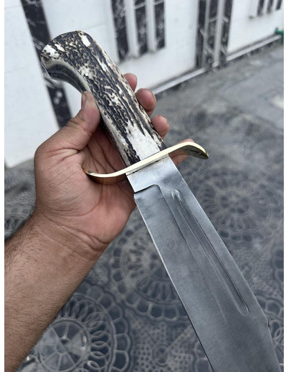 Large handmade full tang Bowie knife with brass guard and genuine stag handle displayed outdoors.