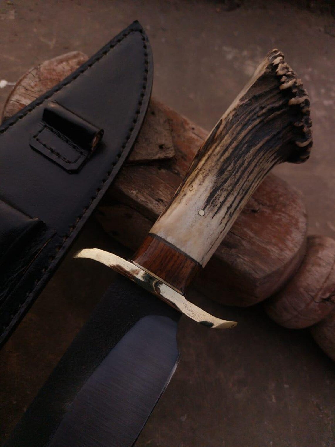Full-length handmade carbon steel Bowie knife with brass guard and stag handle beside leather sheath.